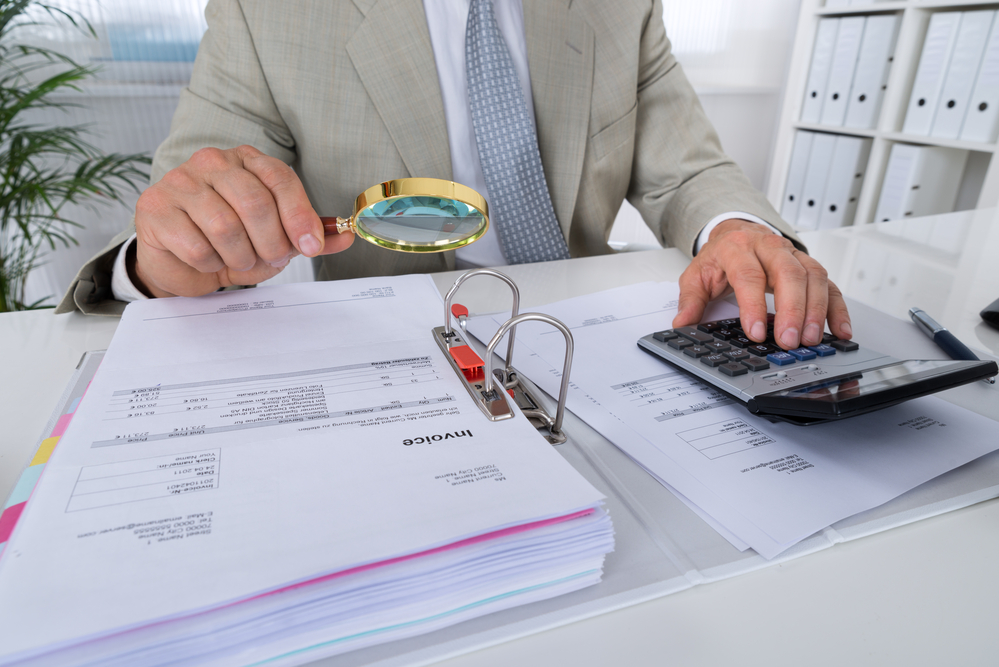 A man holding a magnifying glass over an invoice while using a calculator