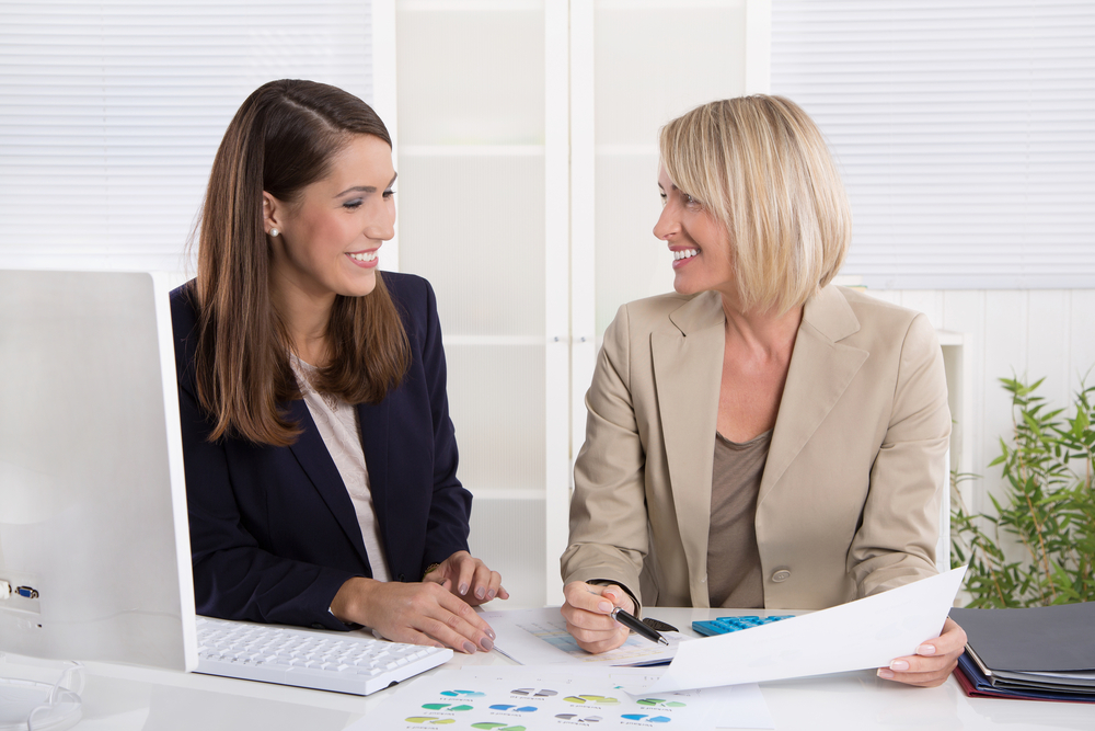 A woman in her 40s training a woman in her late 20s in front of a desktop computer at a modern medical office desk.