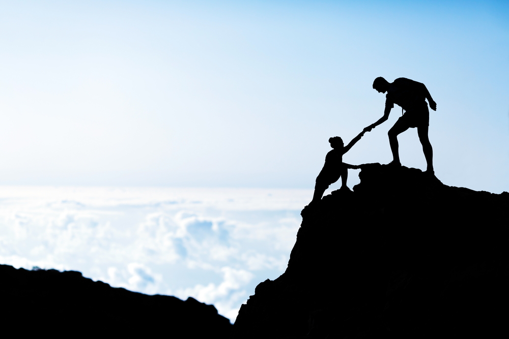 An individual helping another person reach the top of a mountain