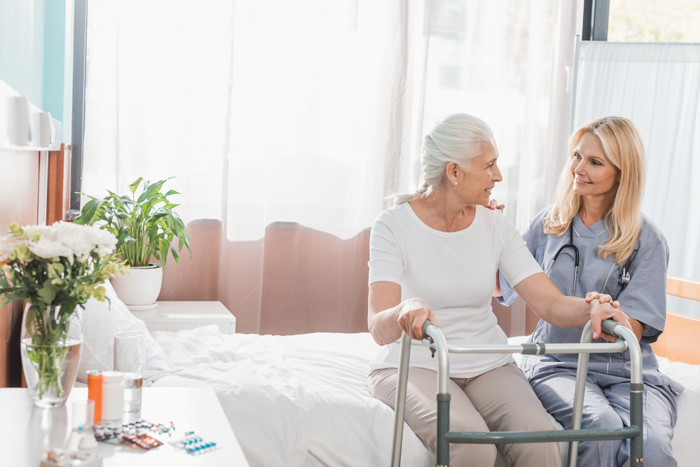 A senior citizen in a nursing home sitting on a bed with a nurse and a walker in front of her.